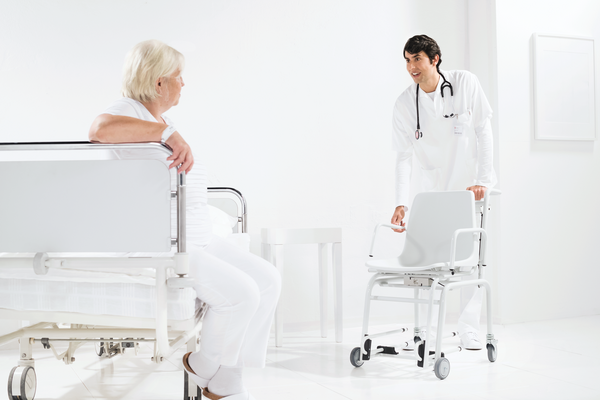 A male nurse holding a SECA chair and an elderly patient woman in a white clinic.