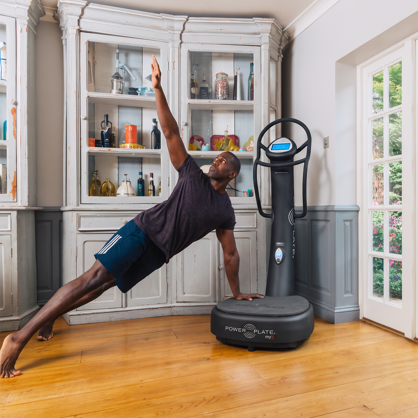 A man in a side plank exercise pose on a wooden floor next to a Power Plate Vibration Stability machine in a bright room with white French doors and a large cabinet.